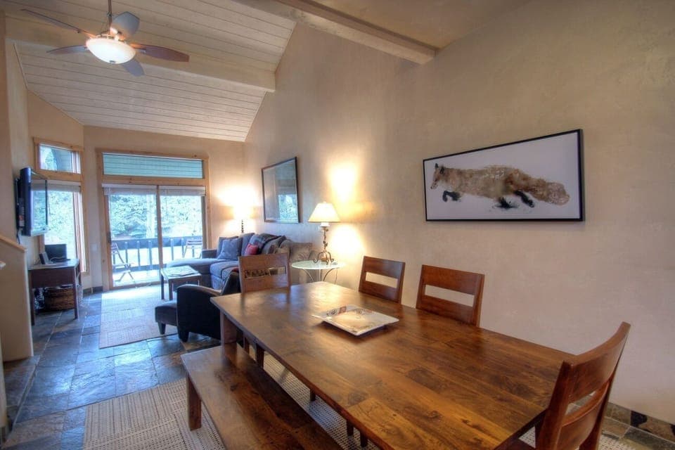 A cozy living and dining area with a wooden table, bench seating, and a couch. The room has a vaulted ceiling, ceiling fan, and large windows leading to a balcony. Artwork hangs on the wall.