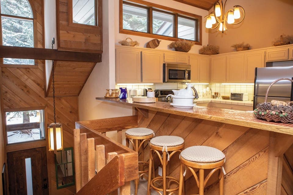 A cozy kitchen with wooden accents, three bar stools at a counter, white cabinets, and stainless steel appliances. Stairs lead down beside the kitchen. Large windows provide natural light.