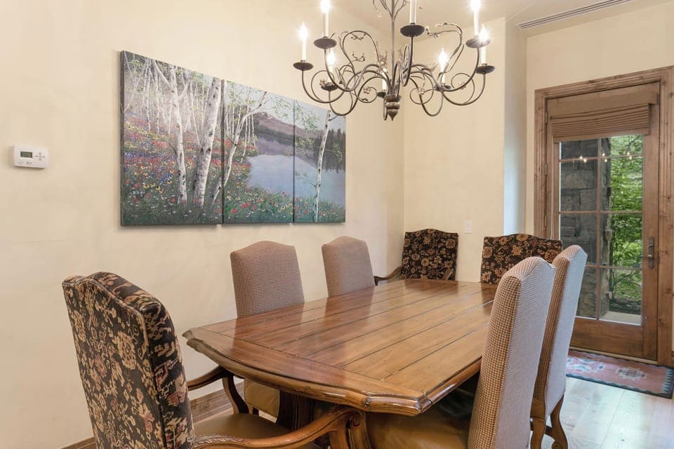 A dining room with a wooden table, eight chairs, a chandelier, a large landscape painting on the wall, and a glass door leading outside.