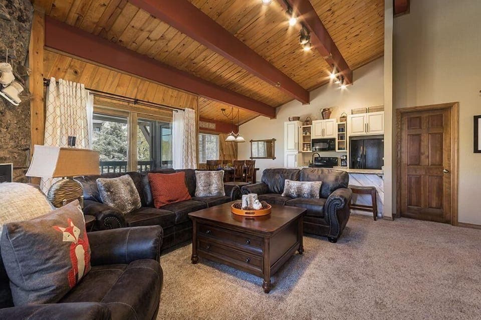 A cozy living room features dark leather sofas, a wooden coffee table, a vaulted wooden ceiling with beams, and a view into a kitchen and dining area. Natural light enters through large windows.