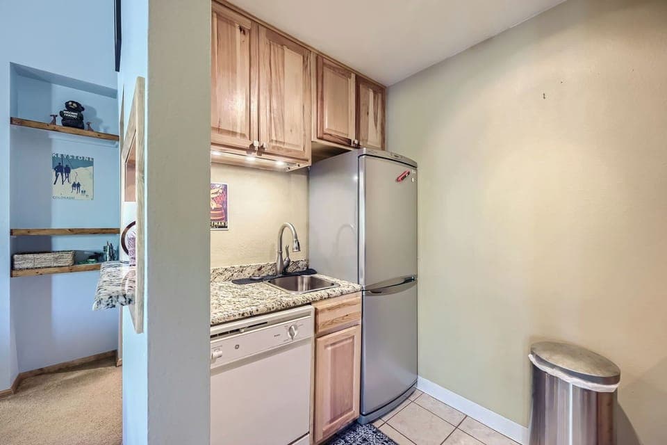 Compact kitchen with wooden cabinets, granite countertops, stainless steel sink, and fridge. Dishwasher below the counter. White tile floor and a silver trash can in the corner.