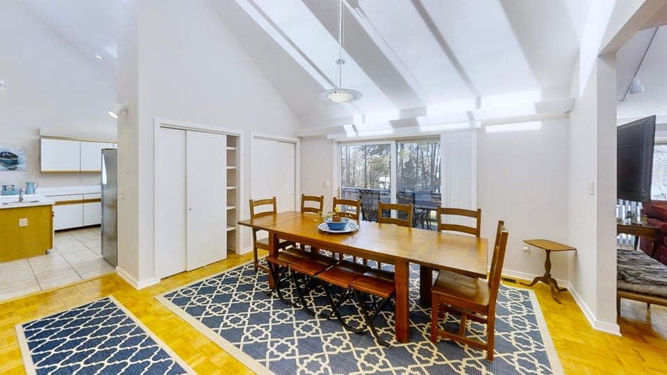 A dining area with a wooden table set for eight overlooking a deck and lake through sliding glass doors.
