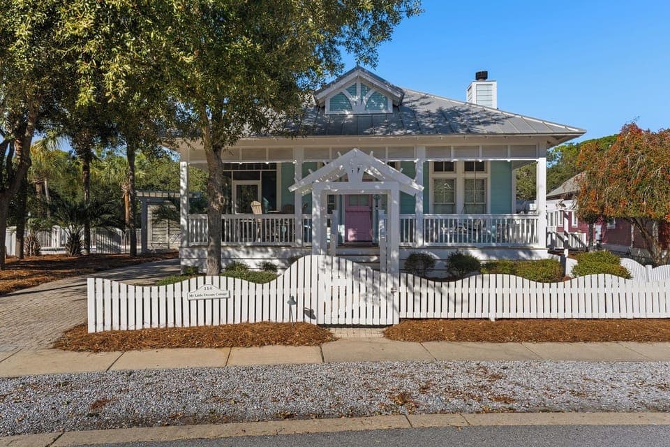 Charming coastal cottage with wraparound porch and picket fence, nestled among mature trees in a peaceful neighborhood setting.