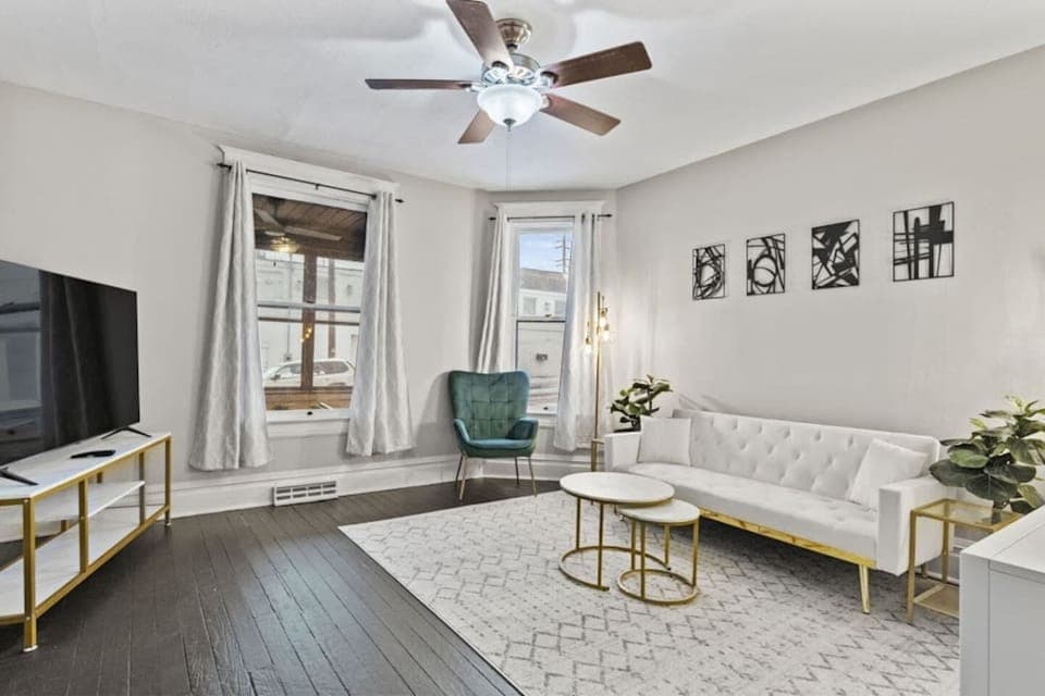 Bright living room featuring a large window with stylish curtains, a chandelier ceiling fan, an elegant sofa, tasteful decorations, mini side tables, and a polished, sophisticated floor design—perfectly blending comfort and modern elegance