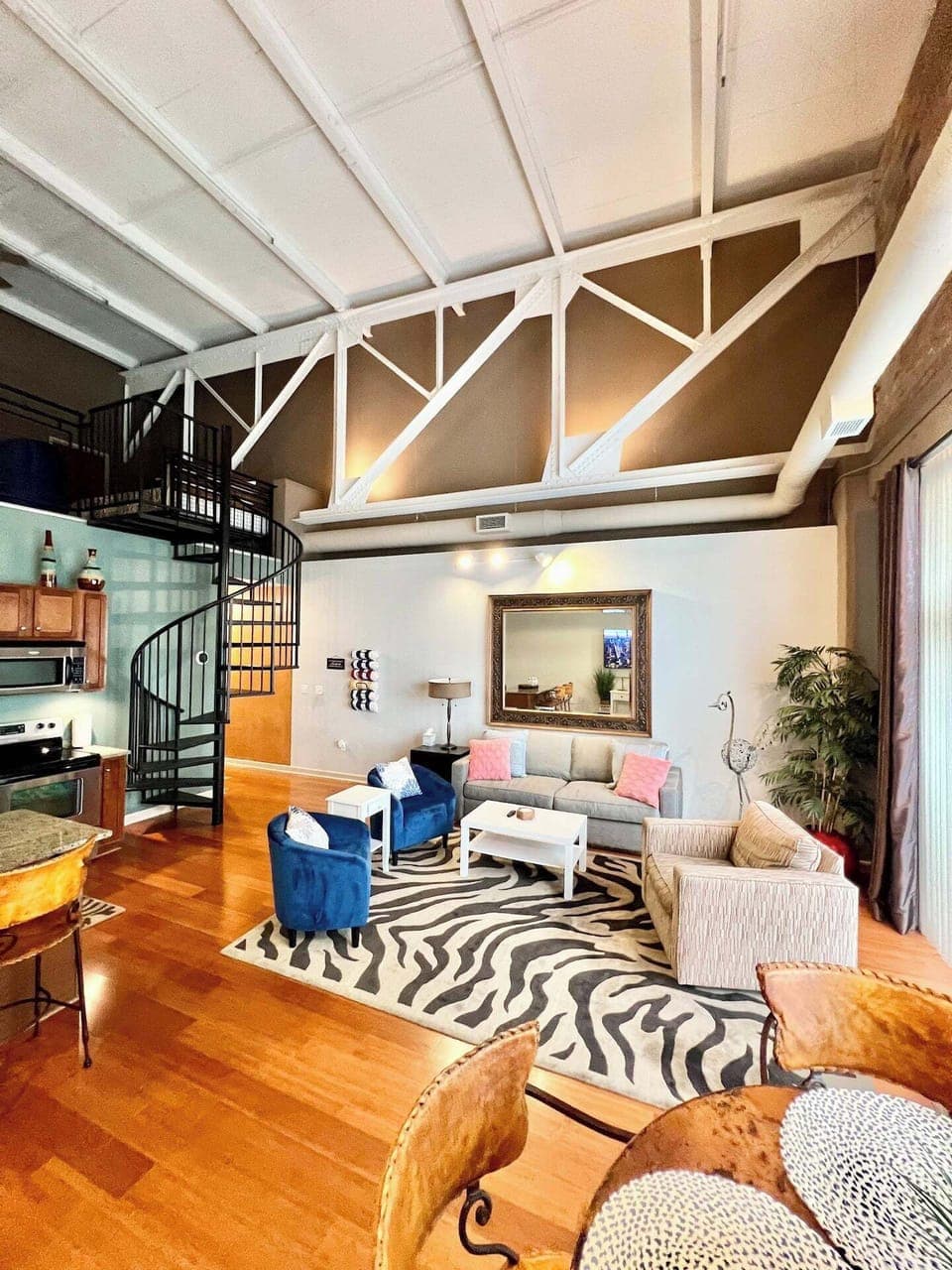 This vibrant living area features vaulted ceilings with exposed beams, a spiral staircase, and colorful accents. The open-concept design includes a modern kitchen, cozy seating, and a zebra-patterned rug for a bold yet welcoming ambiance.