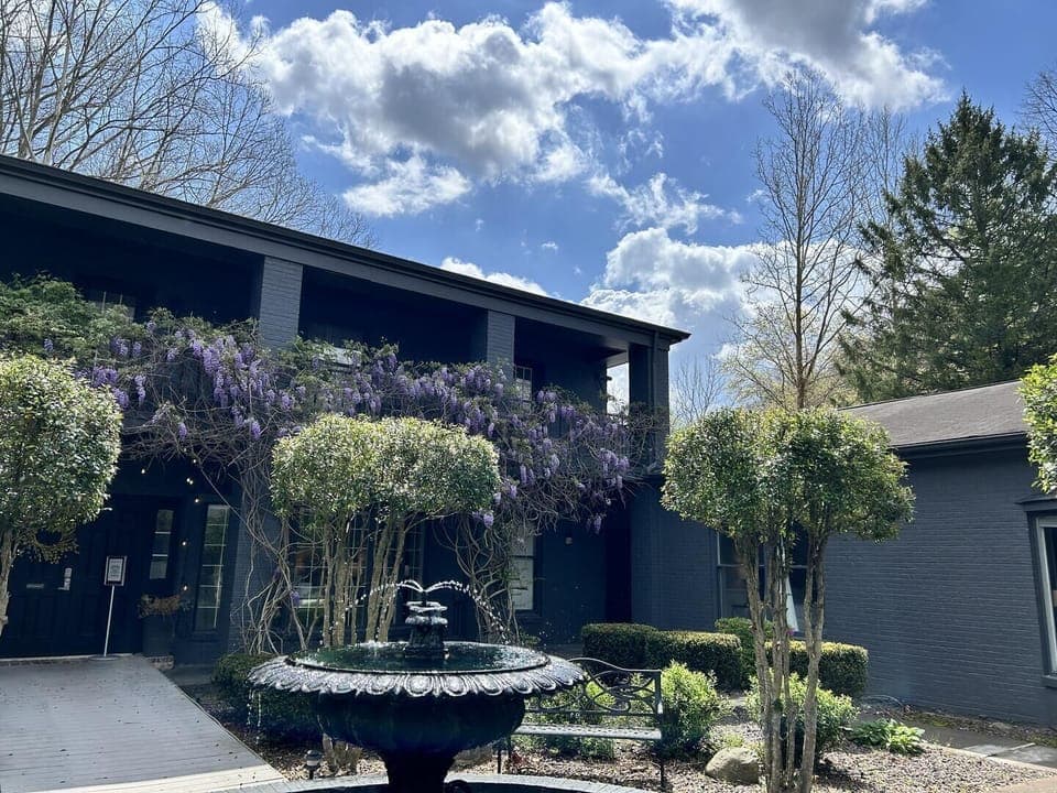 Black brick exterior with purple wisteria vines, manicured shrubs, and a tiered water fountain in front courtyard.