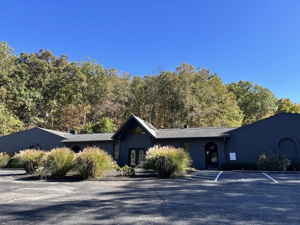 Exterior view of charcoal-gray building with triangular glass entry, surrounded by ornamental grasses and trees.