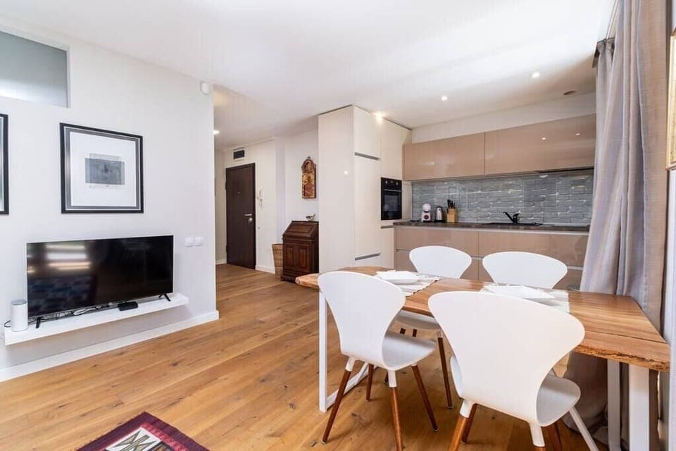 Open-concept space with wooden floors, a sleek kitchen with neutral cabinets and a tiled backsplash, a dining table with white chairs, and a cozy TV area. Bright and modern, offering a comfortable and stylish atmosphere for guests.