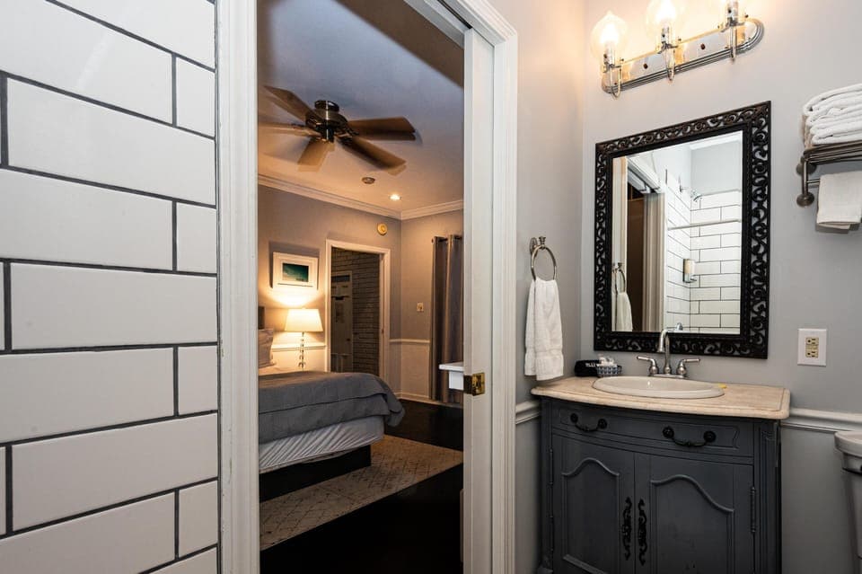 Bathroom sink with ornate black-framed mirror and marble countertop, view into bedroom with gray bedding and ceiling fan.