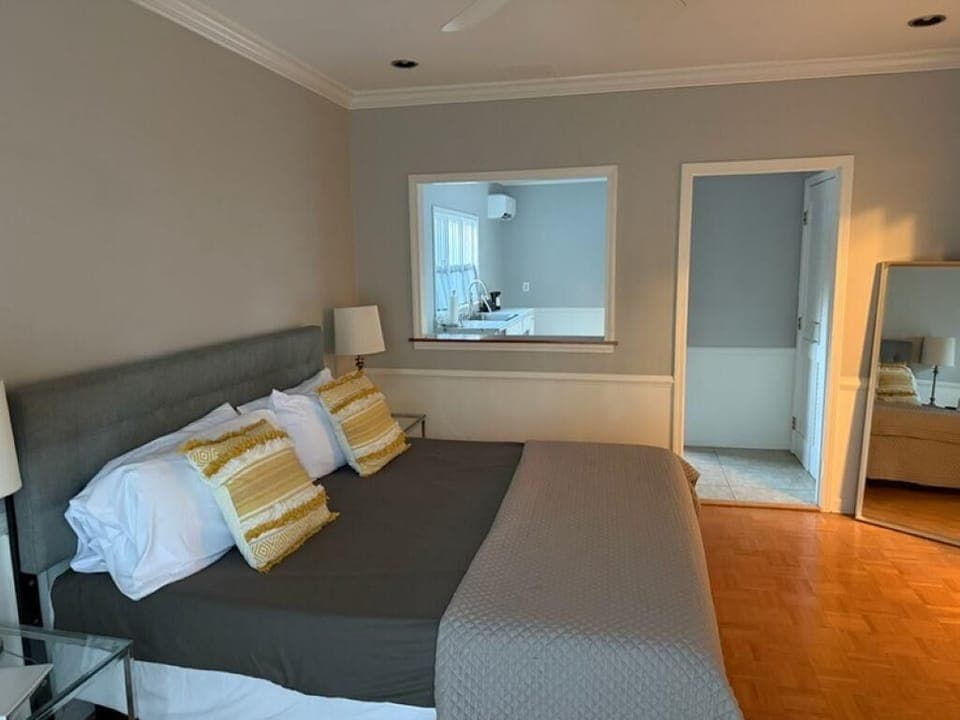 King bed with gray headboard, yellow patterned pillows, and a view into the white kitchen through an interior window.