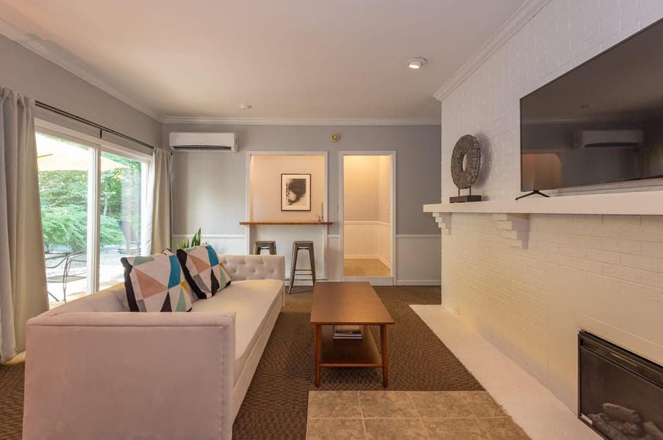 Living area with white tufted sofa, geometric pillows, wood coffee table, and wall-mounted TV over painted brick hearth.