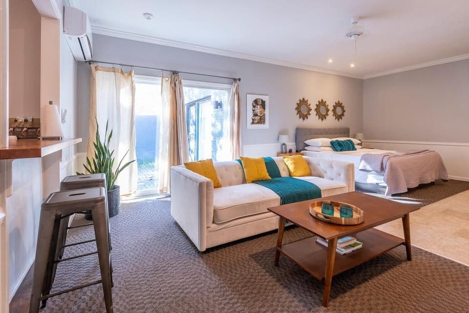 Open-concept suite with beige tufted sofa, wooden coffee table, queen bed, and sheer curtains over sliding glass door.