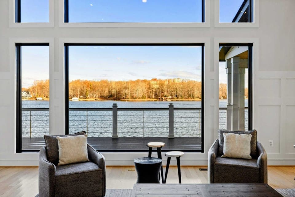 This stunning great room features soaring windows with breathtaking lake views, plush sectional seating, a modern fireplace, and seamless access to the kitchen and dining areas, creating the perfect space for relaxation and entertaining.