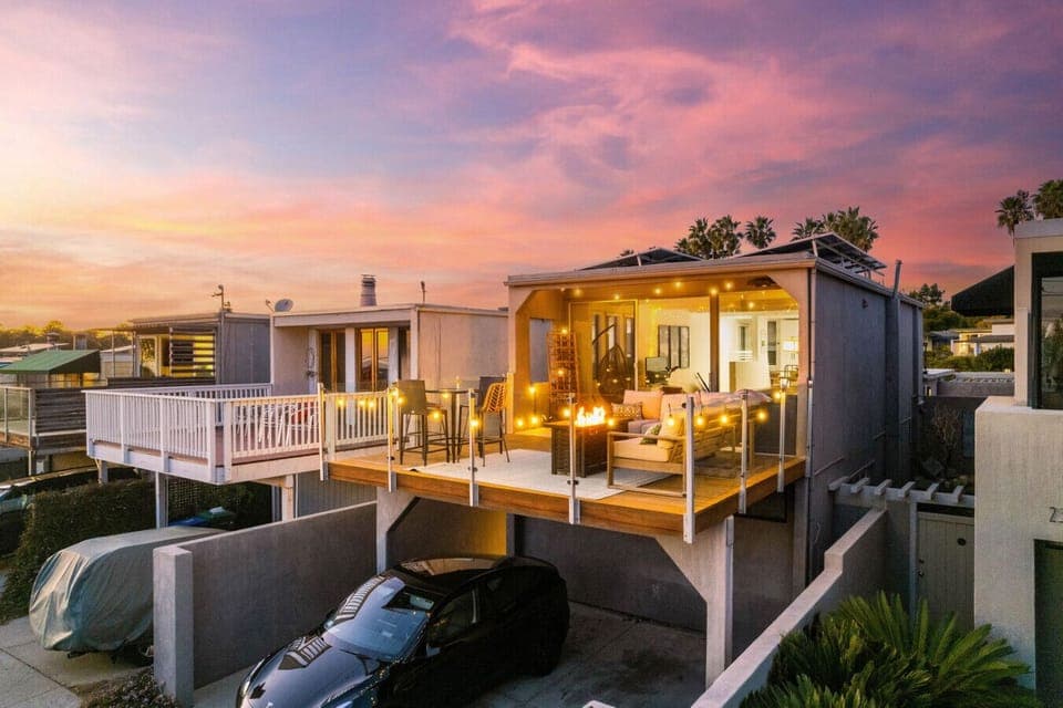 The house features a spacious outdoor deck lit by cozy string lights, providing a warm ambiance. The open sliding doors reveal a well-lit and inviting interior, seamlessly blending indoor and outdoor living spaces.