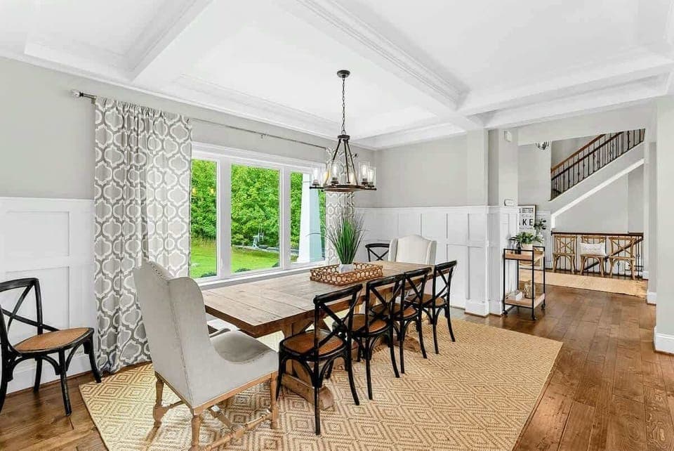 A bright and elegant dining room with a large farmhouse table, stylish seating, coffered ceilings, and a window offering scenic views of the backyard.