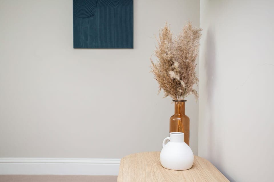 A minimalistic display with a vase of pampas grass and a sleek white jug on a wooden table, complemented by abstract artwork on the wall.