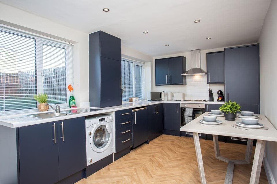 This image shows a bright kitchen with navy blue cabinets, modern appliances, and a small dining table with place settings. The kitchen has natural light streaming in through the window.