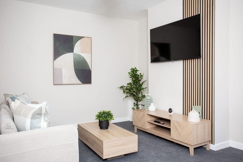 A cozy living room with a soft sofa, a TV mounted on the wall, a simple wooden coffee table, and a decorative potted plant.