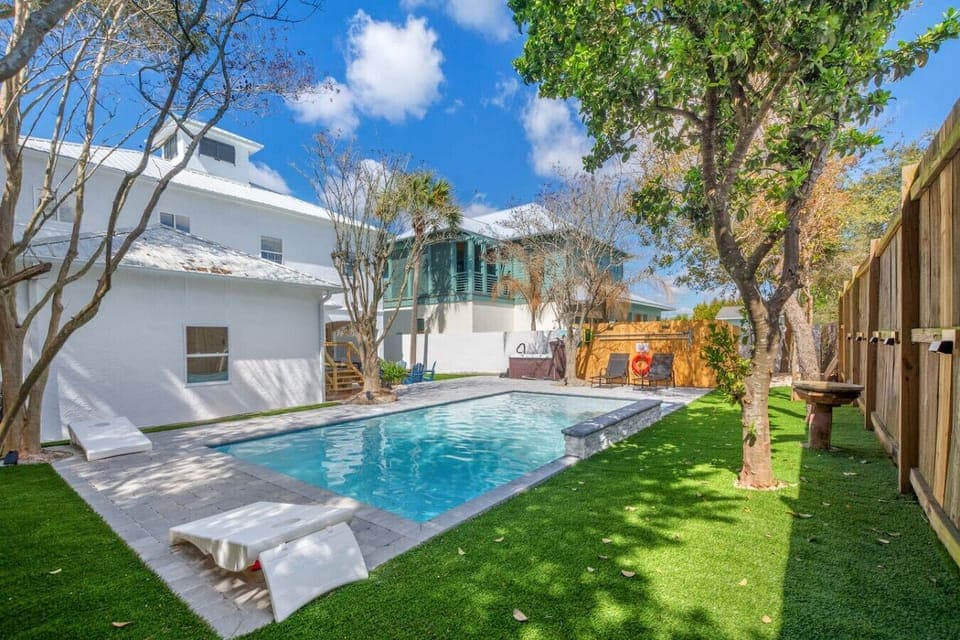 A spacious backyard with a pool, hot tub, fenced-in yard, trees, and a fun corn hole setup!
