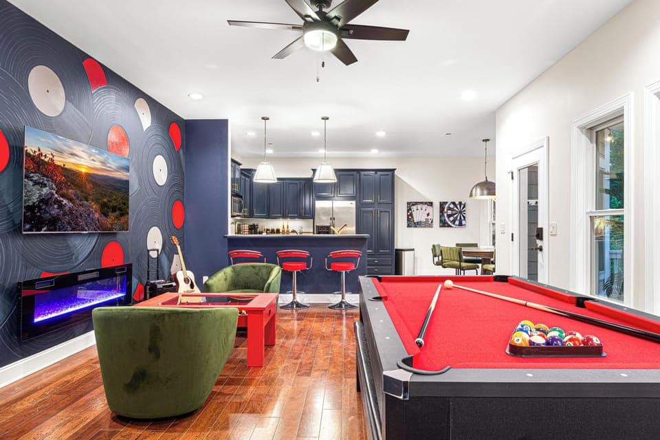 A vibrant entertainment space featuring a red-felt pool table, a cozy seating area with green velvet chairs, and a modern electric fireplace beneath a mounted TV. The stylish bar area, adorned with navy blue cabinetry and red barstools, seamlessly connects to a game corner with a dartboard and poker artwork, creating the ultimate game room experience.