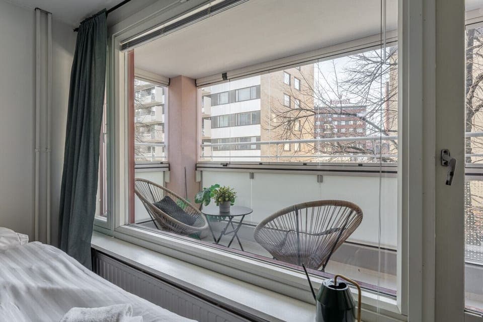 The balcony features comfortable rattan chairs and a small table for outdoor lounging. Enjoy a quiet moment with a view of the surrounding city trees.