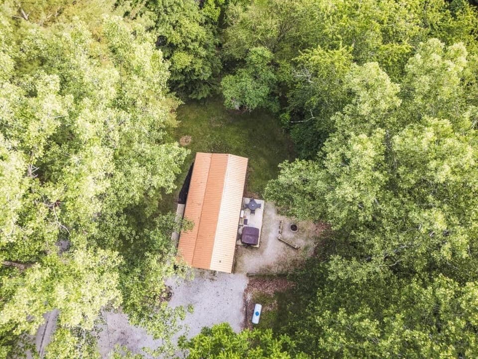 Aerial view of a cabin surrounded by lush green trees, featuring a gravel driveway, outdoor patio with furniture, and a fire pit area — perfect for a serene nature getaway 