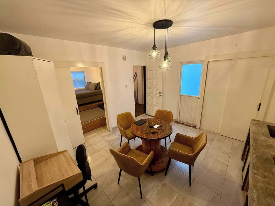 Sala vista desde la esquina(mesa con 4 sillas, escritorio individual con silla, lámpara de lectura, cocina integrada, espejo alto, clóset, barra con estufa integrada, tarja, repisas con tazas, copas y licores, refrigerador, stand con snacks, minibar)