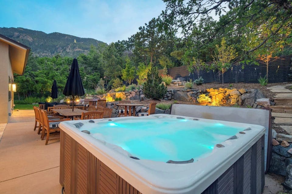 Steaming hot tub set into a stone patio, glowing under soft evening lights with outdoor dining tables nearby and wooded hills all around—an inviting spot to sink in, unwind, and watch the sky fade into dusk.