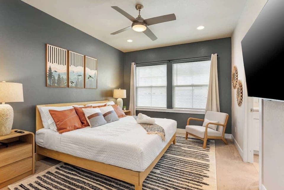 Bright and inviting bedroom with a king-sized wooden bed, white and burnt orange bedding, and matching pillows. Features two bedside lamps, a cozy armchair, a ceiling fan, a large window for natural light, and a wall-mounted TV for your comfort.