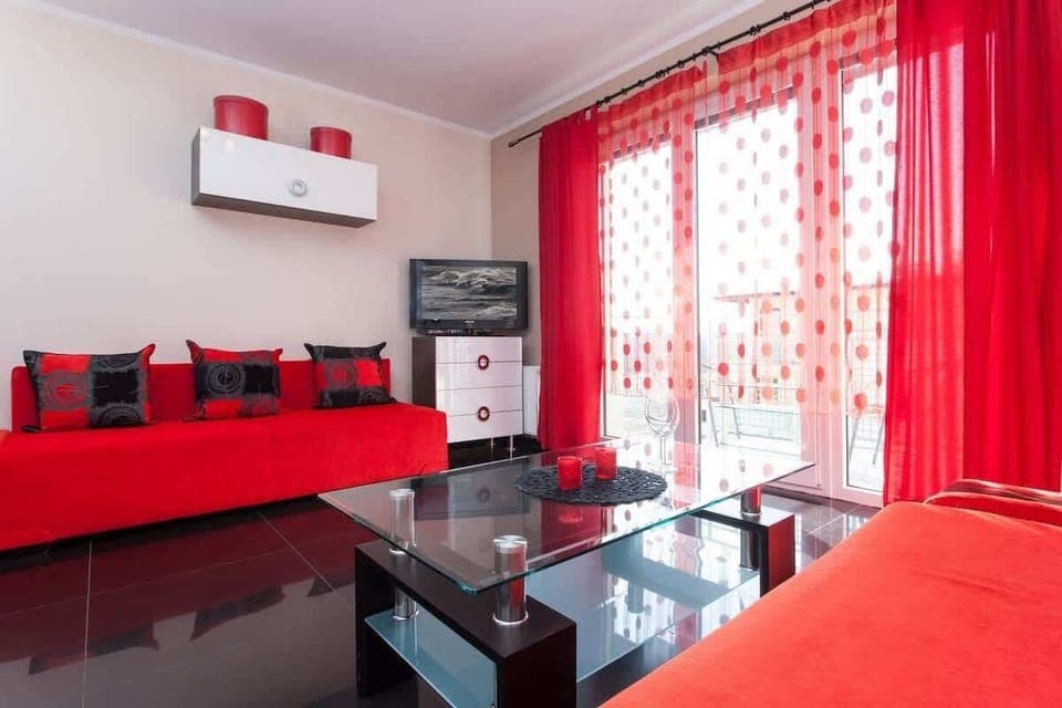 A vibrant living space featuring a red sofa, matching curtains, and decorative pillows. The glass coffee table and black flooring add a modern touch.