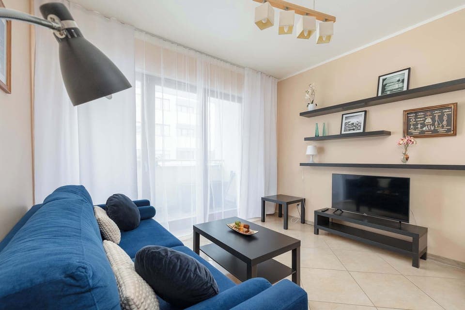 A bright room with a blue sofa, coffee table, and a large window. A TV is visible in the background.