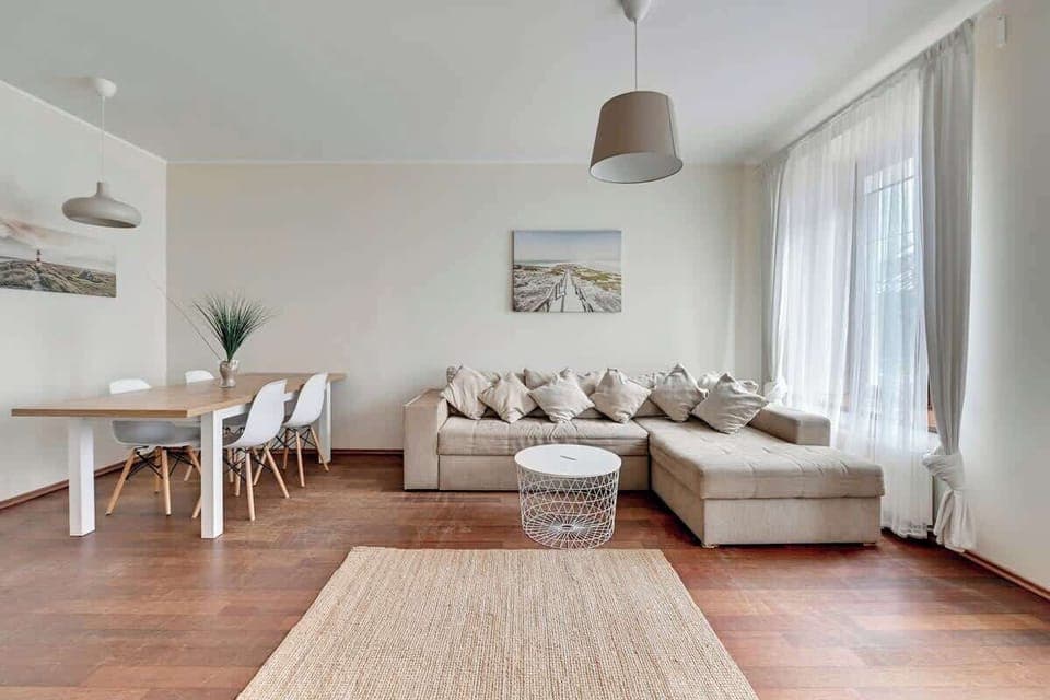 A stylish and minimalist living room with a soft sofa, a small coffee table, and a modern rug. The space is filled with natural light from the large windows, creating a warm and inviting atmosphere.