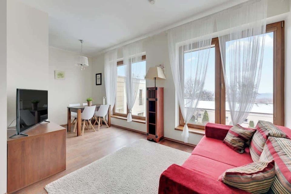 A modern living room featuring a red sofa and a simple coffee table, bathed in natural light coming through large windows, creating a warm and inviting space.
