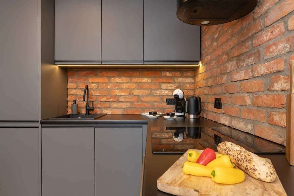 A compact kitchen with grey cabinets, brick backsplash, and a fruit bowl on the counter.


