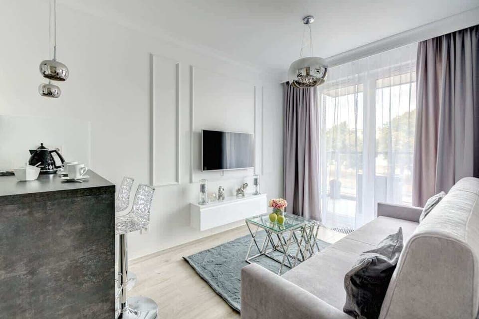 An elegant living room with a light gray sofa, a glass coffee table, decorative wall paneling, a mounted TV, and floor-to-ceiling windows with sheer and heavy curtains, creating a bright yet sophisticated atmosphere.