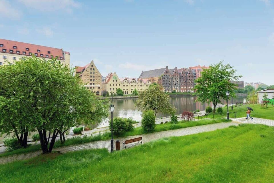 Picturesque riverside scene showcases a charming blend of lush greenery, historic buildings with distinctive gabled roofs, and tranquil pathways, evoking a serene, timeless atmosphere.