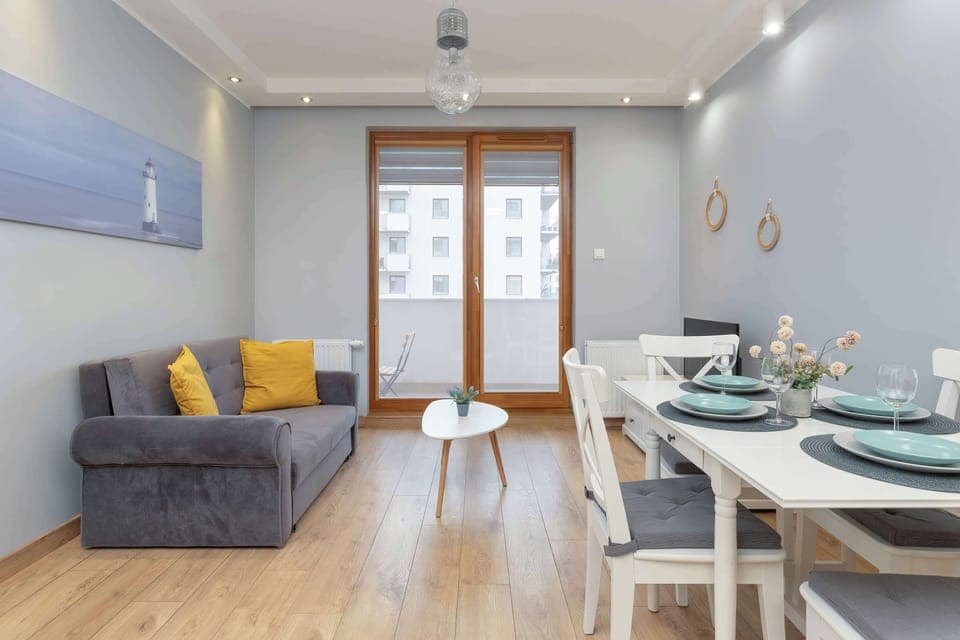 A bright living room with a sofa, coffee table, and a television unit. Yellow and grey decor create a modern and cozy ambiance. White dinning table.