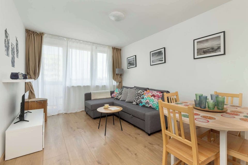 A bright living room with a gray corner sofa, TV, dining table, and large window letting in natural light.