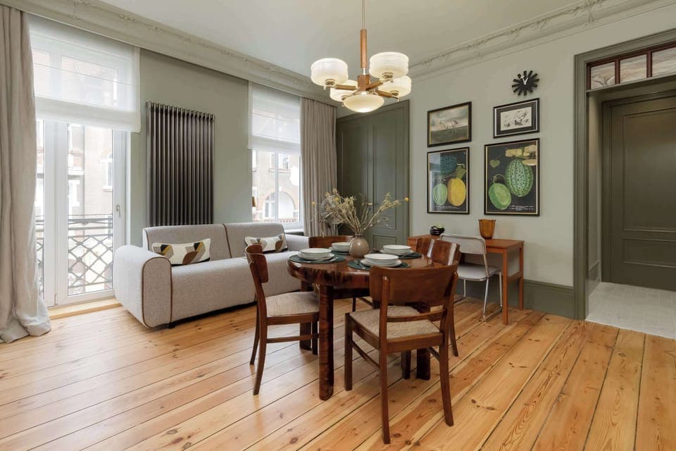 A dining and living area with a round wooden table, elegant chairs, and large windows letting in natural light.