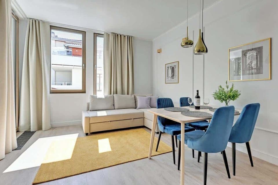 A well-lit living room with a modern sofa, a small coffee table, and dining chairs beside a large window.