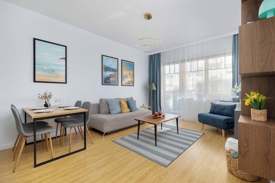 Bright living room with modern decor, wooden floor, blue sofa, wall art, and large windows letting in natural light.