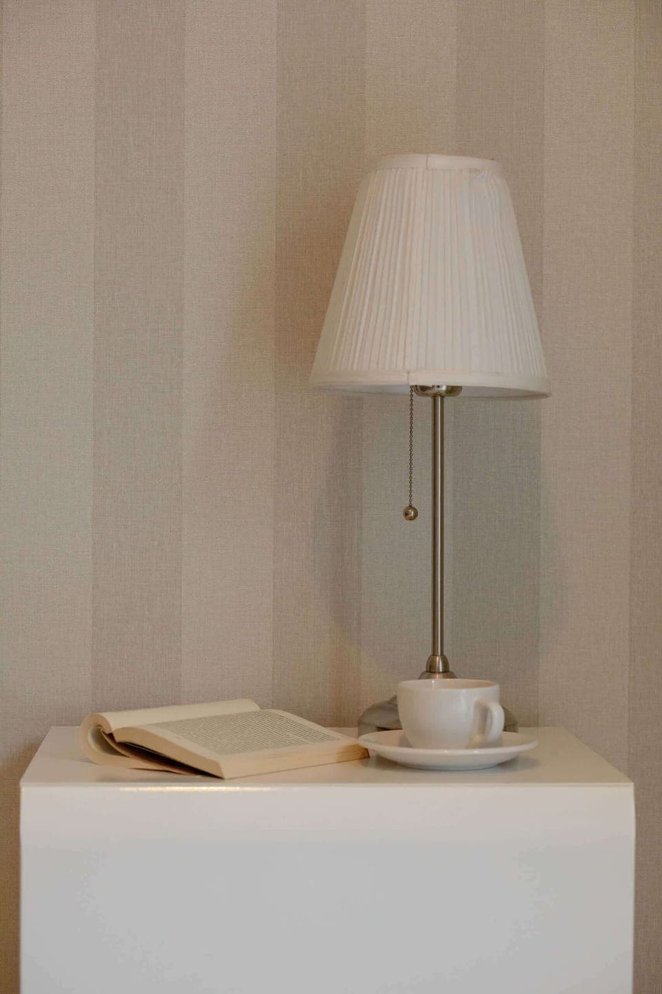 A close-up of a bedside table with a modern white lamp and a neatly arranged book, creating a cozy nightstand setup.


