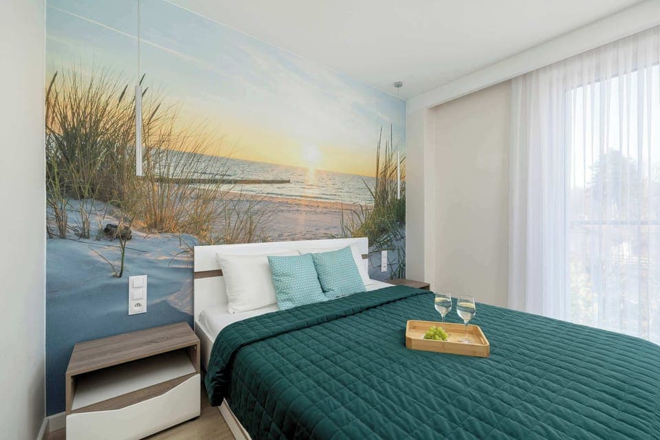 A modern bedroom in the apartment at Morska 12/23 in Rewal, equipped with a large bed with a turquoise bedspread and pillows, two bedside tables with lamps and a decorative wall mural will allow you to quickly regenerate after a long day.