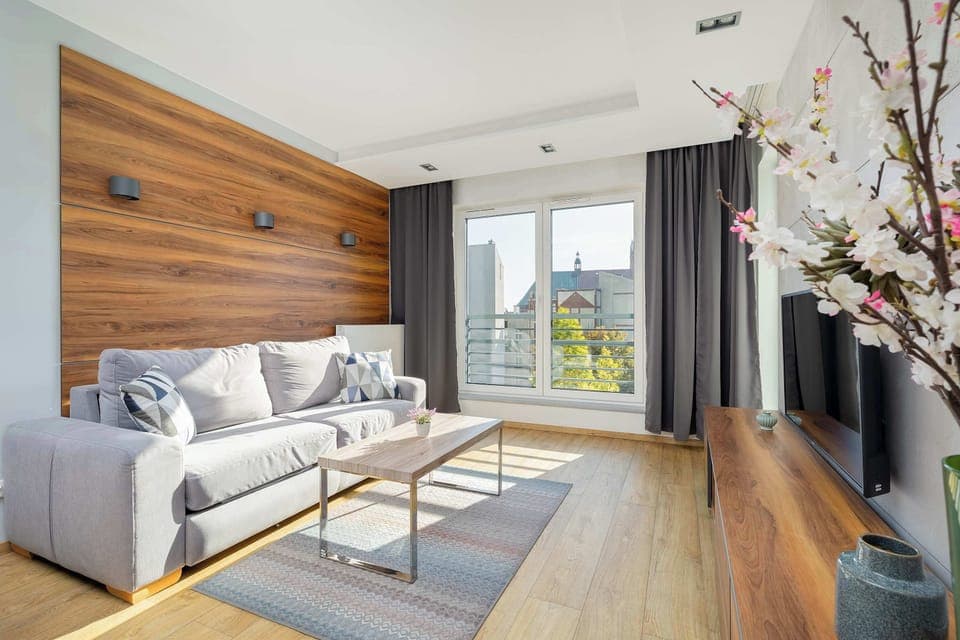 A bright living room with modern decor, including a grey couch, a light wood coffee table, and a flat-screen TV. Large windows allow natural light to brighten the space, and the decor has a soft, neutral palette.
