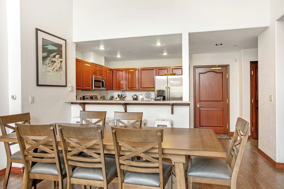 A kitchen and dining area featuring wooden cabinets, a long wooden dining table with chairs, bar stools at a counter, and a framed picture on the wall.