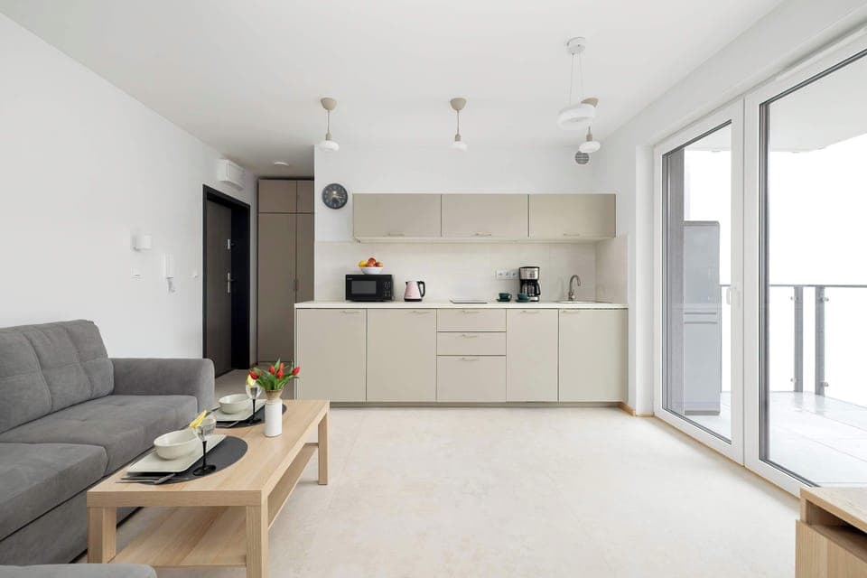 A cozy open-plan living room with a gray sectional sofa, light walls, and a compact kitchen area in soft beige tones.