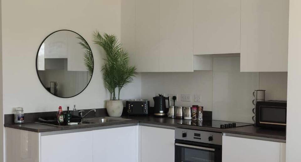 Stylish, fully equipped kitchen with modern appliances including oven, microwave, kettle, and toaster. Sleek white units, black worktops, and a round mirror create a clean, contemporary feel perfect for home-cooked meals during your stay.