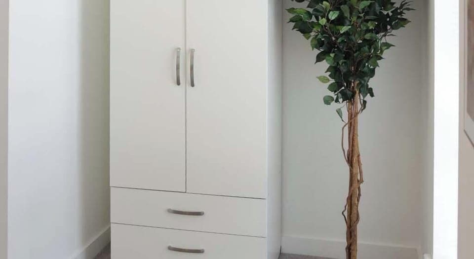Simple white wardrobe with two doors and two drawers, providing ample storage space. Positioned next to a tall, leafy artificial plant, adding a touch of greenery and a fresh feel to the room’s calm, neutral decor.