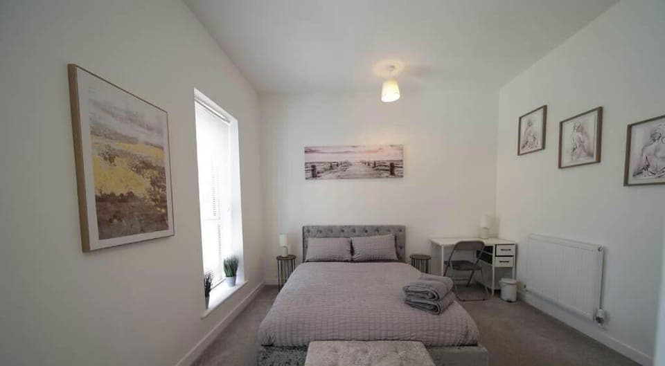 Light and airy bedroom with a cosy double bed dressed in soft grey bedding. Towels are neatly folded on the bed. The room includes a small desk, ottoman, radiator, and framed artworks on the walls, creating a calm and welcoming space.