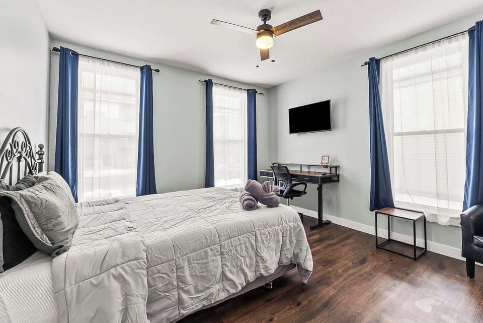 Bright bedroom view showcasing large windows, blackout curtains, ceiling fan, and a dedicated work desk.